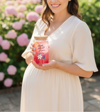 Pregnant woman holding a pink jar with floral design and text, standing outdoors with flowers in the background.