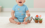 Baby wearing a blue onesie with gardening tools and plants on a light background