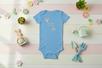 Blue baby onesie with floral design on a colorful wooden surface with toys and plants.