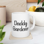 White mug with 'Daddy Gardener' text on a wooden coaster, surrounded by plants and a bowl of fruit.