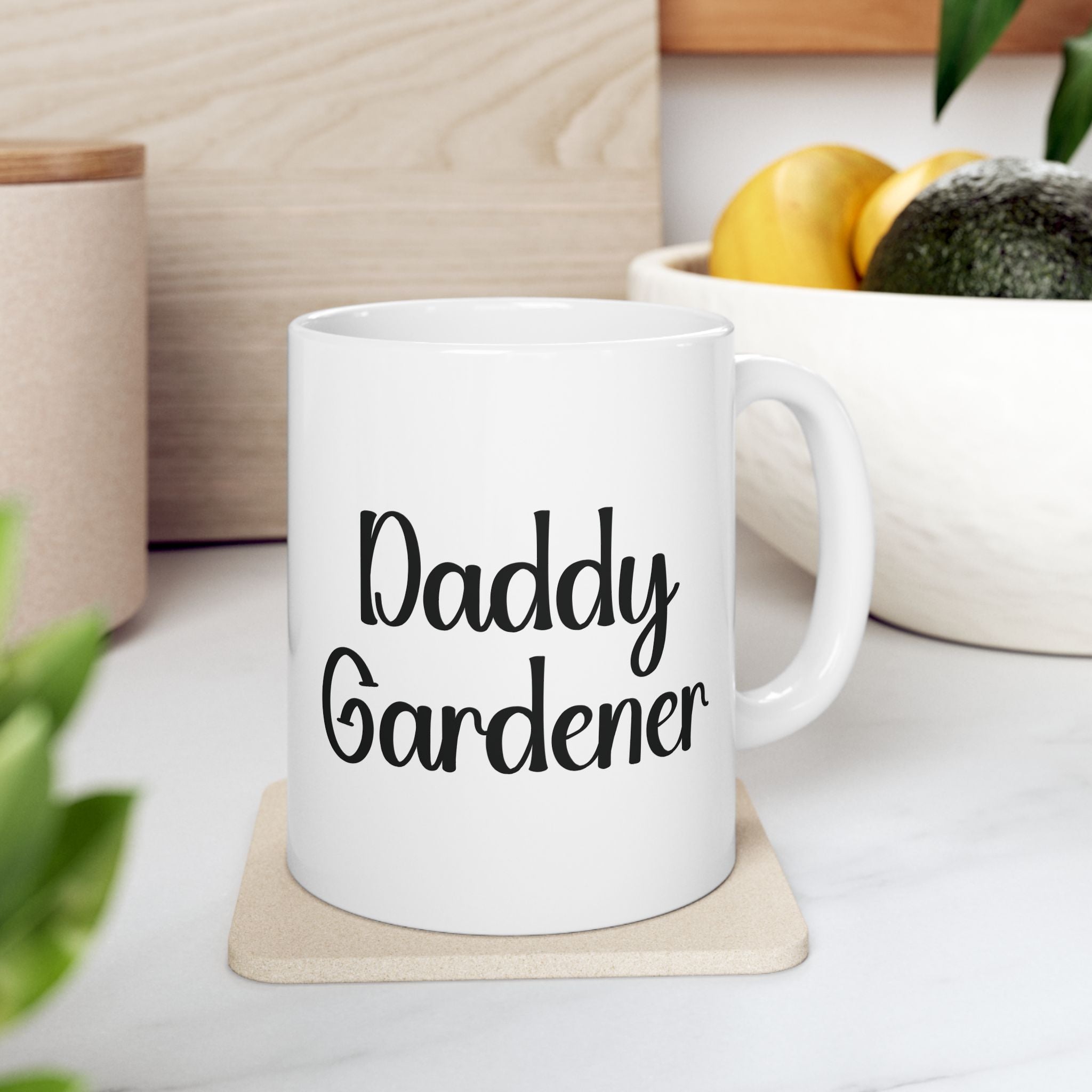 White mug with 'Daddy Gardener' text on a wooden coaster, surrounded by plants and a bowl of fruit.
