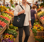 A man in a market aisle carrying a black tote bag with a colorful 'Fresh Good!' graphic. The background shows a bustling outdoor farmers market with produce displays.