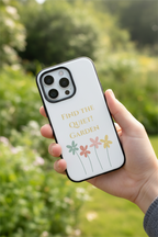 Hand holding a phone case with floral design and text 'Find the Quiet Garden' against a blurred green background.