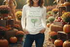 Woman wearing a white sweatshirt with a tree graphic and text, standing in an autumn-themed setting with pumpkins and flowers.