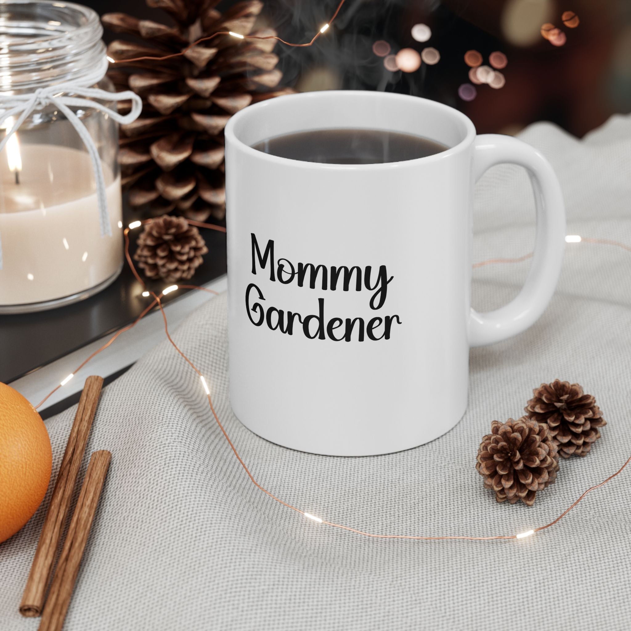 White mug with 'Mommy Gardener' text on a table with decorative items like pinecones and candles.