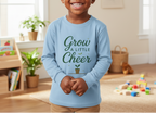 Child wearing a blue shirt with 'Grow a Little Cheer' text in a room with toys and plants.