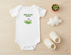 White baby onesie with 'New to the Patch' text and plant graphics on a wooden surface with baby shoes and a small plant.
