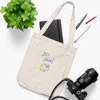 Beige tote bag with 'Every Day Tote' text, camera, notebook, and stationery on a white background
