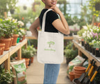 Woman holding a tote bag with 'Garden Strong' logo in a greenhouse setting