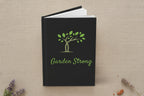 A hardcover notebook or journal with a black cover, featuring a green tree graphic and the words 'Garden Strong' in green script on the front, shown on a white background.