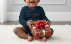 Child wearing a 'Merry Christmas' shirt holding a gift box with a red bow on a white carpet.