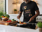 Man cooking in a kitchen wearing a black t-shirt with 'Spring into Gardening' text and floral designs.
