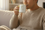 Man in a beige sweater holding a mug with a logo, sitting on a couch.