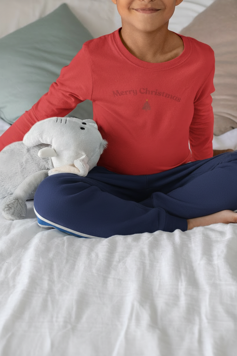 Child wearing a red 'Merry Christmas' shirt sitting on a bed with a gray pillow and white blanket.