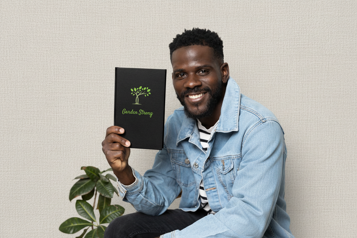A smiling man in a denim jacket holds up a black notebook with a green tree logo and the words 'Garden Strong' on the cover, set against a clean white background with a plant leaf visible in the lower left.