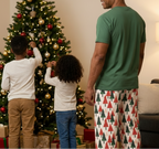 A family of four wearing matching Christmas pajamas stands in a living room in front of a decorated Christmas tree. The father and child on the right wear white long-sleeve tops with a red, green, and white geometric pattern on the back, paired with matching patterned pants.