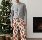 A man stands barefoot in a living room wearing a gray long-sleeve shirt and cream pajama pants with a red and green Christmas tree pattern. A decorated Christmas tree and a lit fireplace are visible in the background.