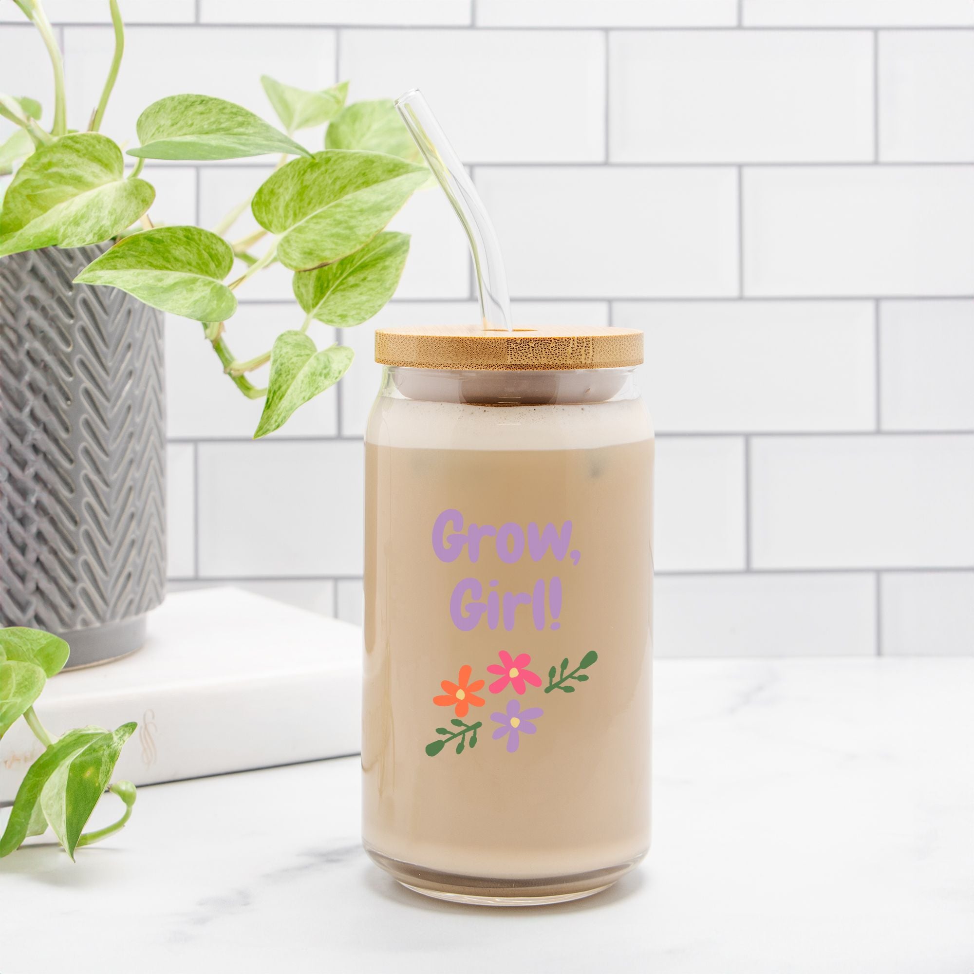 Candle in a glass jar with a wooden lid and colorful text and graphics on a white surface with a plant in the background.