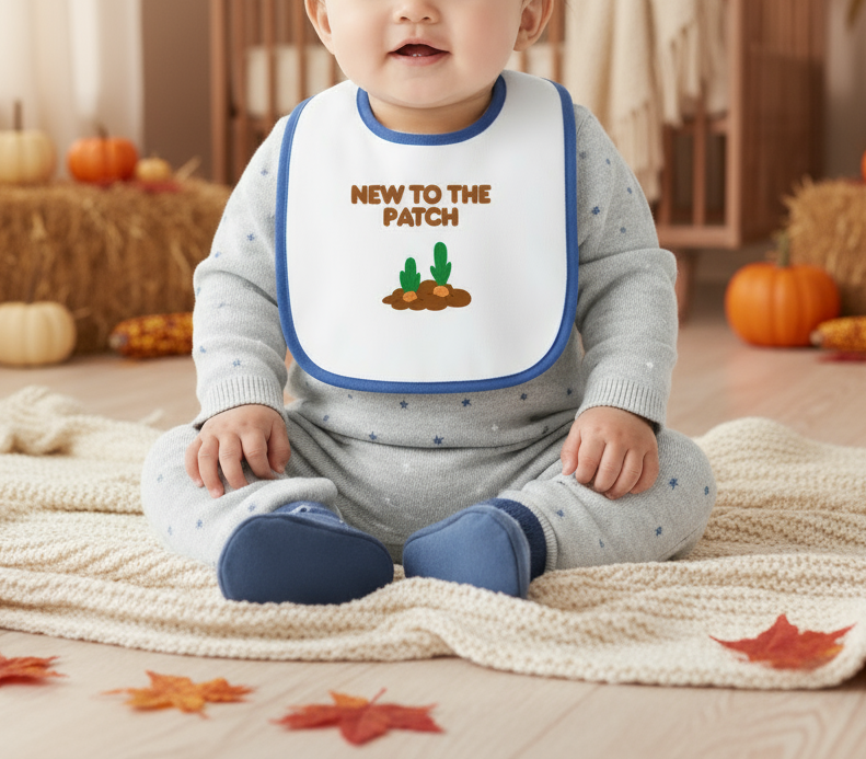 Baby wearing a bib with 'New to the Patch' text and pumpkin graphics, sitting on a blanket with pumpkins and leaves in the background.