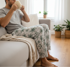 Man sitting on a couch in a living room, holding a mug and wearing pajama pants with a plant pattern.