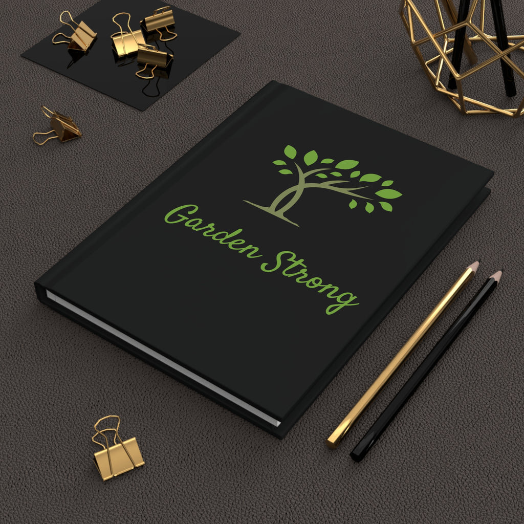 A black hardcover notebook with a green tree logo and the words 'Garden Strong' on the cover, styled on a dark textured surface with gold and black binder clips, pencils, and a geometric decor object.