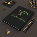 A black hardcover notebook with a green tree logo and the words 'Garden Strong' on the cover, styled on a dark textured surface with gold and black binder clips, pencils, and a geometric decor object.