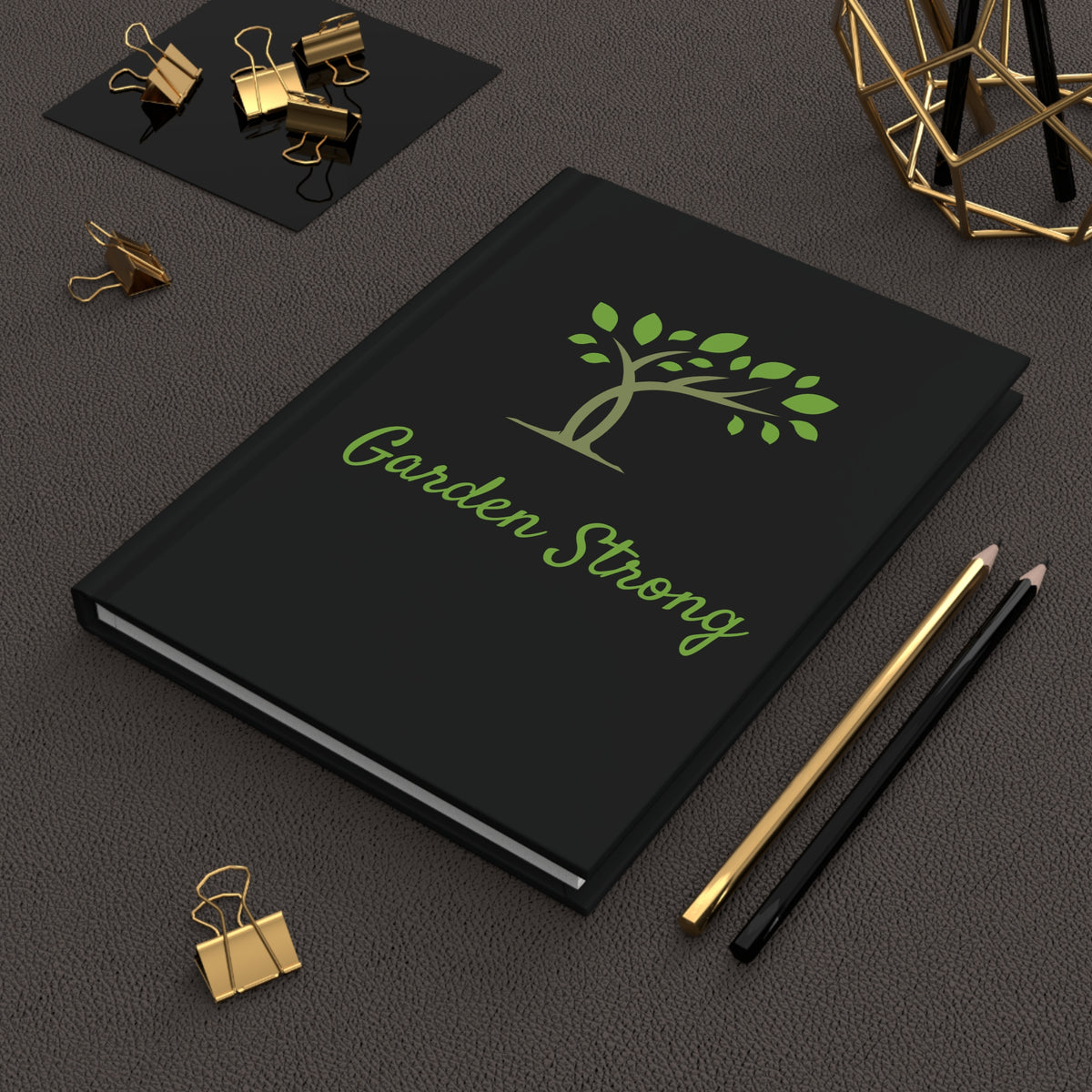 A black hardcover notebook with a green tree logo and the words 'Garden Strong' on the cover, styled on a dark textured surface with gold and black binder clips, pencils, and a geometric decor object.