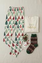 A holiday-themed flat lay featuring a pair of cream pajama pants with a green and red pine tree pattern, a cream folded sweatshirt with gold 'Happy Holidays' lettering, and a pair of green and red patterned knit socks, arranged on a light fabric background.