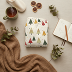 A folded garment or fabric item with a Christmas tree pattern in muted red, green, mustard yellow, and tan on an off-white background, styled in a cozy flat lay with a mug of tea, knit blanket, dried orange slices, eucalyptus leaves, and an open notebook.
