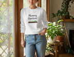 Woman wearing a white sweatshirt with 'Mommy Gardener' text in a home setting with plants.