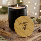 Black candle with wooden lid labeled 'Awakened Scents' on a wooden surface with blurred lights in the background.