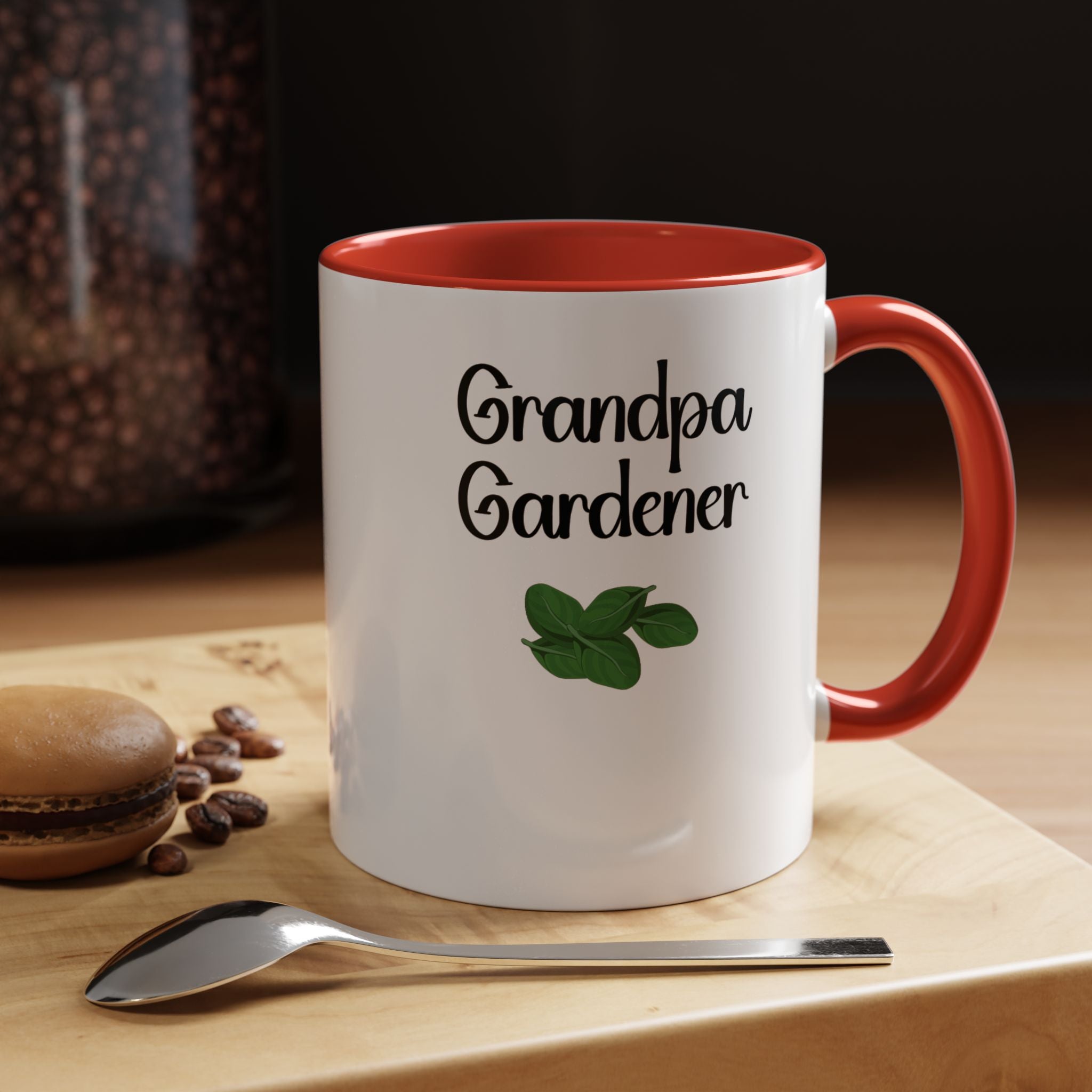 White mug with red interior and handle, featuring 'Grandpa Gardener' text and a leaf graphic, on a wooden surface with coffee beans and a spoon.