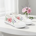 White sneakers with floral designs on a white surface with a vase of flowers.