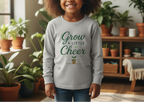 Child wearing a gray sweatshirt with 'Grow a Little Bit of Cheer' text in a home setting with plants.