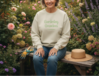 Woman wearing a 'Gardening Grandma' sweatshirt sitting in a garden with flowers and gardening tools.