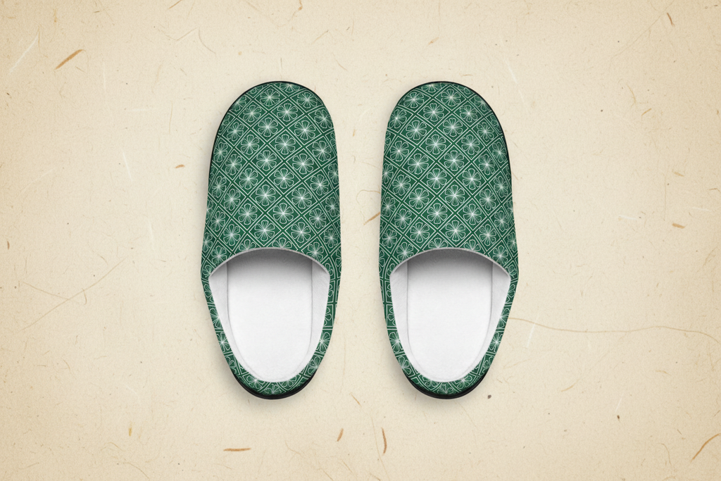 A pair of slip-on house slippers with a green and white geometric pattern, shown from a top-down view on a textured beige background.