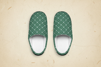 A pair of slip-on house slippers with a green and white geometric pattern, shown from a top-down view on a textured beige background.