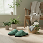 A pair of green slip-on slippers with a small white floral pattern are displayed on a light wood floor in a cozy, sunlit room. The background features a wooden armchair with a draped throw blanket, potted plants, and a basket containing gardening gloves and tools.