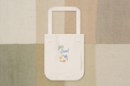 A cream canvas tote bag with white handles, featuring a colorful graphic that reads 'Let's Grow!' in green and blue script with small yellow, blue, pink, and purple flowers. The bag is centered on a textured patchwork background of beige, tan, and sage green linen-like panels.