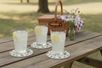 Three glasses of lemonade with lemon slices on a picnic table with a basket and flowers in the background.