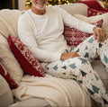 A pair of cream-colored lounge pants featuring a festive Christmas tree pattern with red, green, and blue trees, stars, and snowflakes, displayed flat against a light beige background.