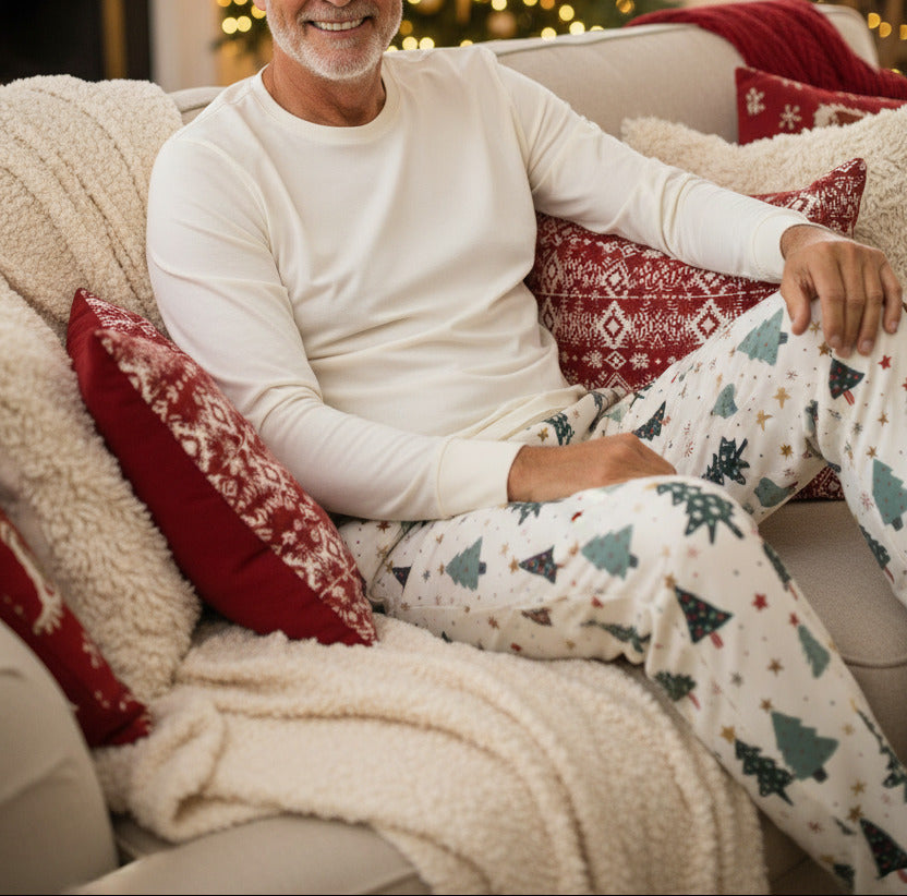 A pair of cream-colored lounge pants featuring a festive Christmas tree pattern with red, green, and blue trees, stars, and snowflakes, displayed flat against a light beige background.
