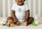 Baby sitting in a crib wearing a white onesie with text, surrounded by toys and a bottle.