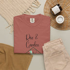 Folded pink t-shirt with 'Rise & Garden' text on a neutral surface with pants and decor items.