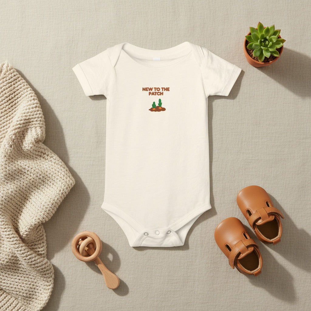 White baby onesie with 'New to the Patch' text and graphic on a beige background