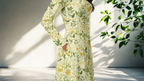 Woman wearing a floral dress with a butterfly design, standing against a white wall with green leaves.