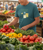 An older man wearing a teal t-shirt with a 'GARDENING GRANDPA' graphic stands at a vibrant outdoor farmers market stall filled with fresh vegetables like red tomatoes, yellow bell peppers, and green kale.