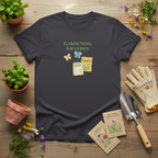 A dark charcoal gray t-shirt with a 'Gardening Grandpa' graphic, styled in a flat-lay on a wooden surface with gardening props including gloves, a trowel, seed packets, and potted plants.