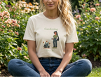 A woman sitting cross-legged in a garden wearing a cream-colored graphic t-shirt with a green and brown illustration of a gardener and the words 'Gardening Try It'.
