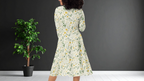 Woman wearing a floral dress standing next to a potted plant against a dark wall.